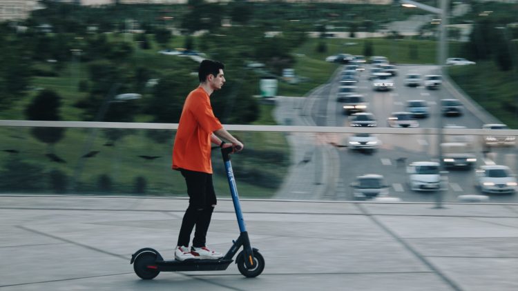 a guy in an orange T-shirt rides an electric scooter over a bridge