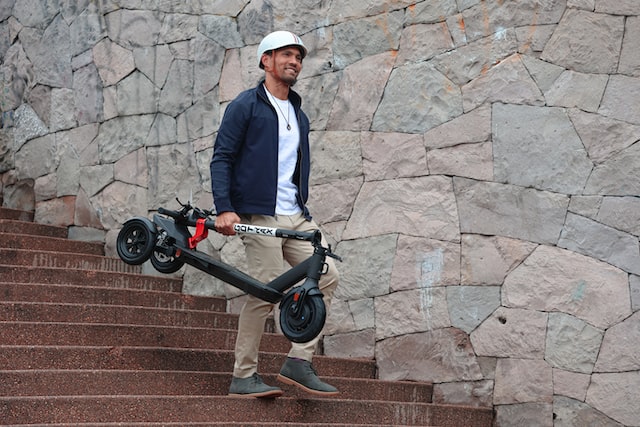 a man in a white helmet comes down the stairs and holds a folded electric scooter in his hand